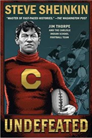 A drawing of a man who is Jim Thorpe looks out to the distance from the cover. He is wearing a red jersey with a large C on it and holding a football.